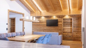 Um interior moderno com teto de madeira, uma televisão de tela plana grande fixada na parede, um banco de madeira com almofadas estampadas e um sofá com almofadas azuis.