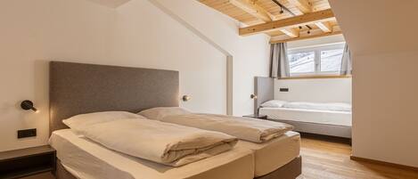 A bedroom with a large bed, a bedside table, a nightstand, a window with curtains, and a sloped ceiling with exposed beams.