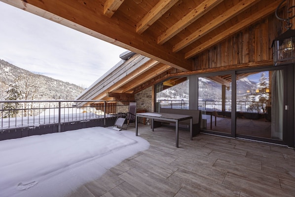 A wooden deck with a table and chairs, a snow-covered ground, and a mountain view through large glass doors.