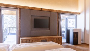Quarto com uma televisão de tela plana grande fixada em uma parede revestida de madeira, um banco de madeira e uma porta de vidro deslizante que dá para uma varanda com vista para uma paisagem nevada.
