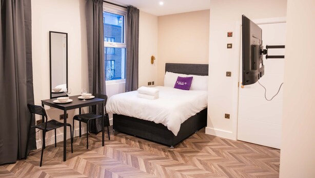 Una camera d'albergo con un letto, una scrivania con sedie, una TV e una finestra con tende.
