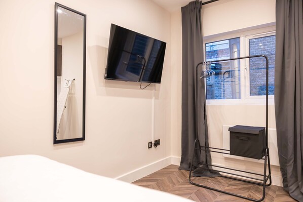 A hotel room with a bed, a TV mounted on the wall, a window with curtains, and a metal rack with a towel. - Spacious Modern Studio in Central London (London)