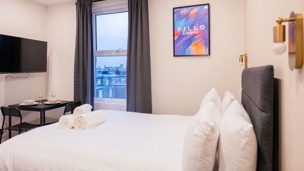 A hotel room with a bed, a desk with a chair, a flat-screen TV, a window with curtains, and a city view. - Modern Studio in Central London (London)