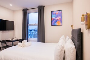 A hotel room with a bed, a desk with a chair, a flat-screen TV, a window with curtains, and a city view.
