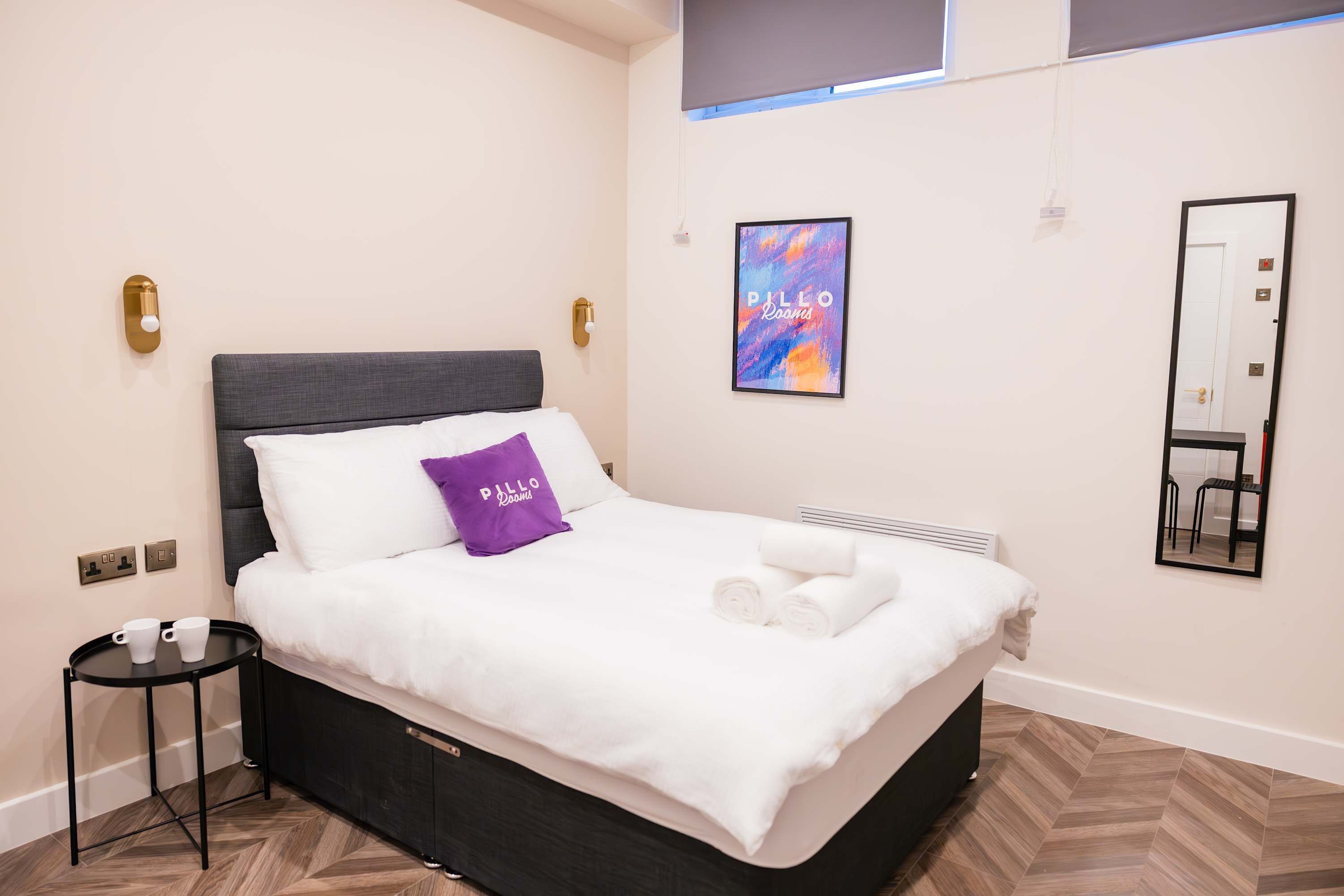 A neatly made bed with a white comforter and a purple pillow, a small side table with two white cups, a framed artwork on the wall, and a mirror.
