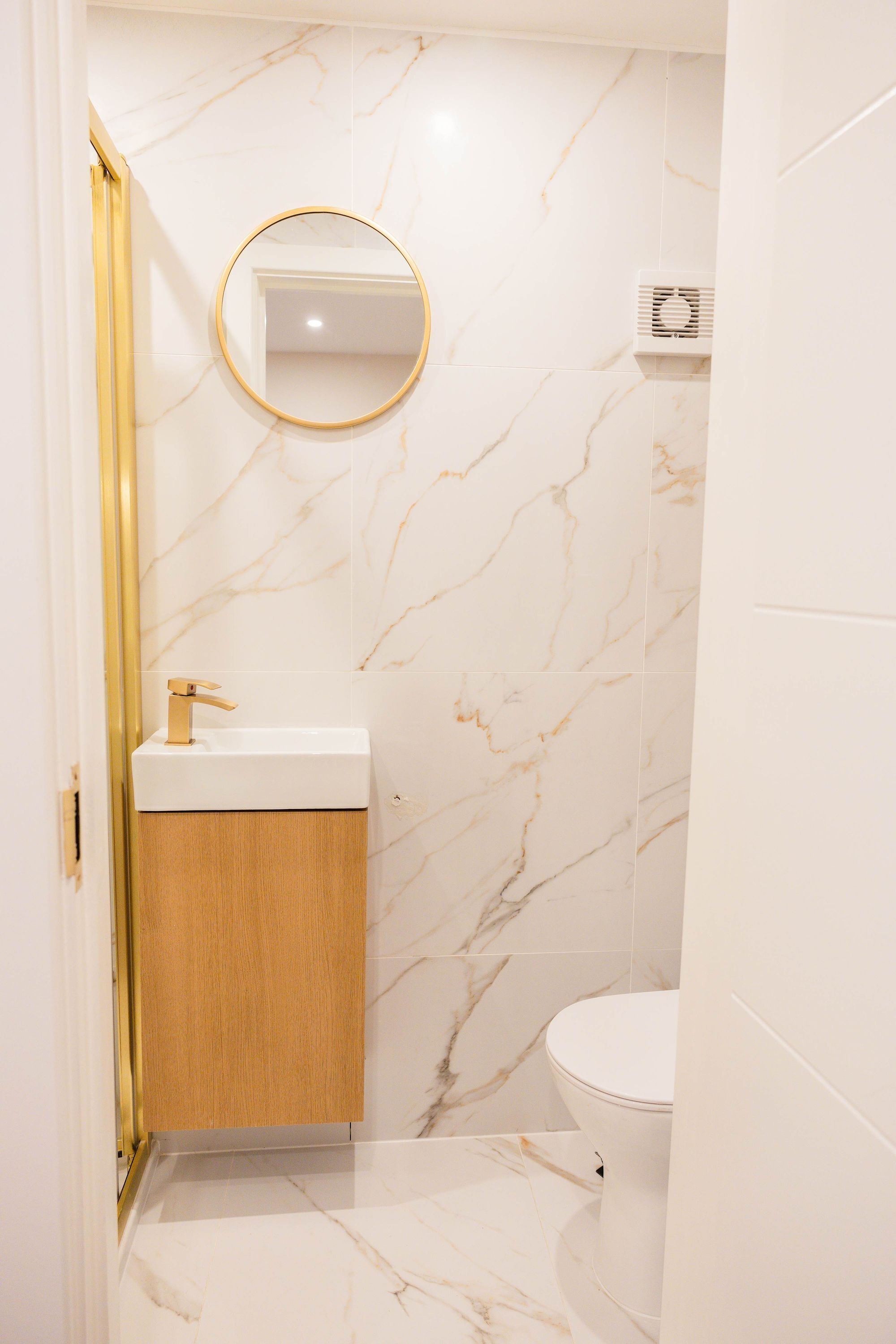 Une salle de bain avec un mur en marbre, un miroir rond, un lavabo blanc et des toilettes.