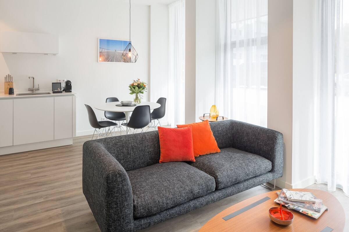 A modern living room with a grey sofa, a wooden coffee table, and a dining area with a white table and black chairs.