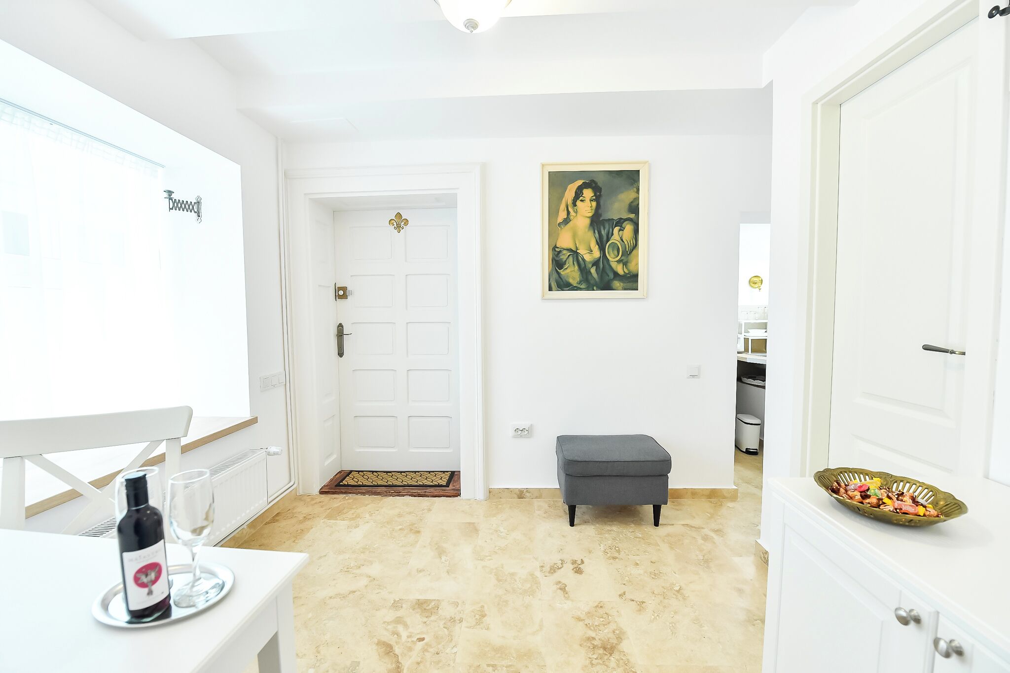A bright, white room with a door, a small gray bench, a white table with a wine bottle and glass, and a painting on the wall.