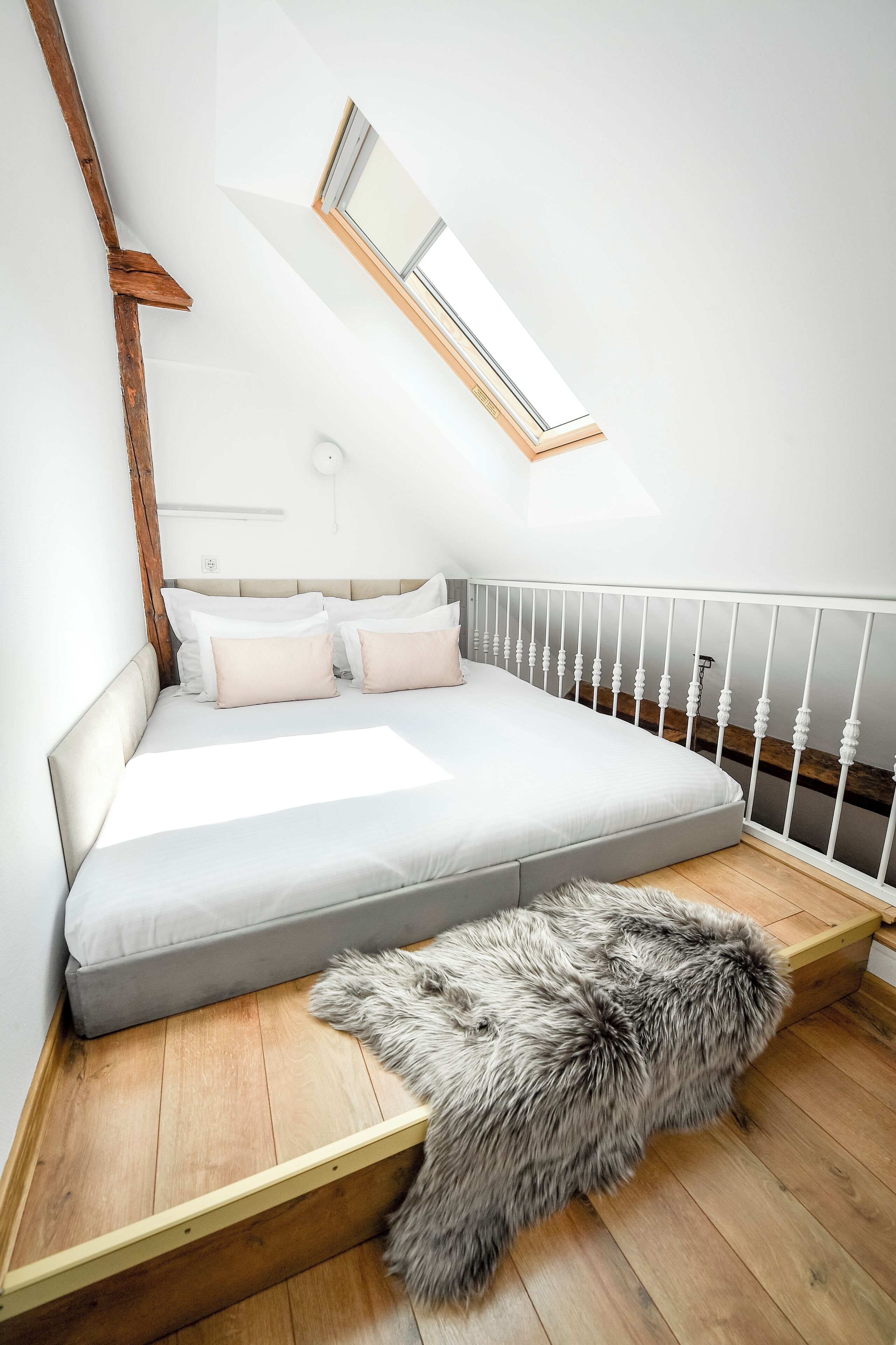 Un dormitorio con techo inclinado, tragaluz, cama con ropa de cama blanca, suelo de madera y manta de piel.