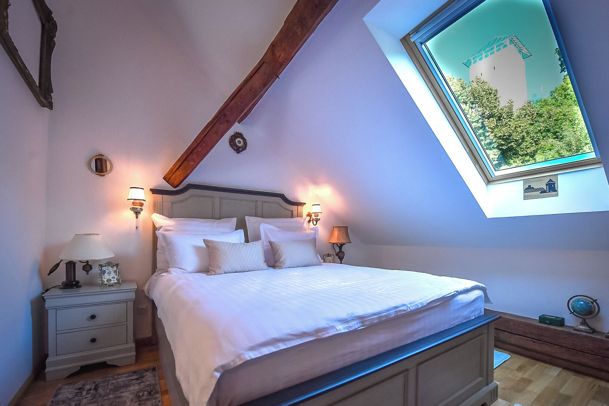 A bedroom with a skylight, a bed with white bedding, two bedside lamps, and a small bedside table with a lamp.