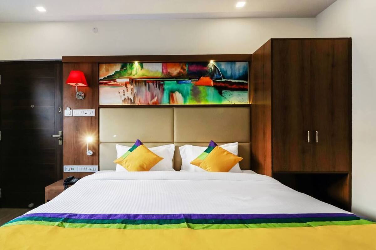 A hotel room with a large bed, a wooden wardrobe, a wall-mounted lamp, and a colorful abstract painting above the bed.