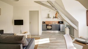Ein modernes Wohnzimmer mit einer Couch, einem Fernseher und einem Essbereich mit Tisch und Stühlen. Im Hintergrund ist eine Küche mit Mikrowelle und Schränken zu sehen.