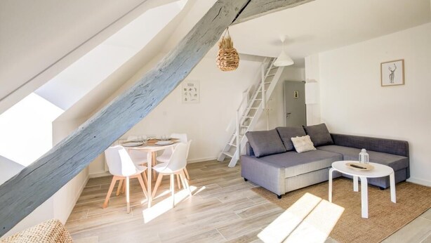 Ein modernes Loft-Apartment mit freiliegenden Holzbalken, einem grauen Sofa, weißen Stühlen und einem Esstisch.