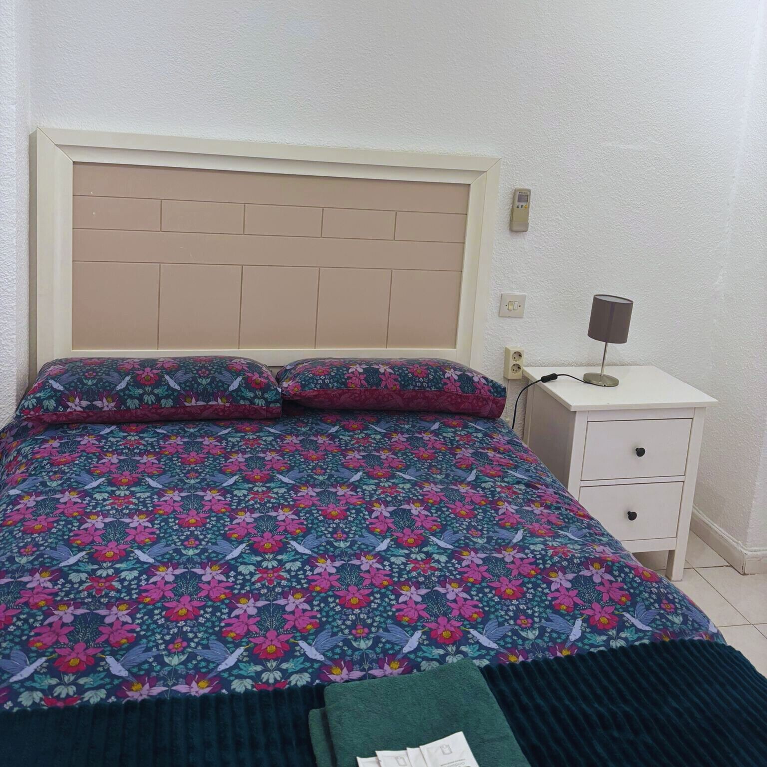 Una cama con edredón y almohadas estampadas con motivos florales, una mesita de noche con lámpara y un enchufe en la pared.