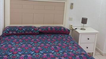 Una cama con edredón y almohadas estampadas con motivos florales, una mesita de noche con lámpara y un enchufe en la pared.