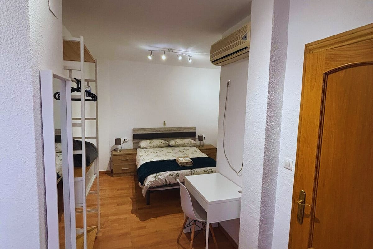 A compact bedroom with a bed, bedside table, and a desk with a chair.