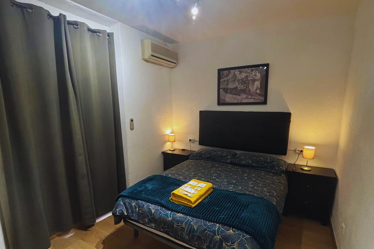 A bedroom with a bed, two bedside tables with lamps, a wall-mounted air conditioner, and a framed picture on the wall.