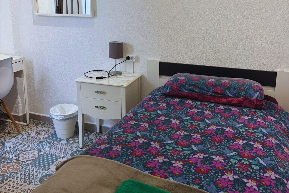 A bedroom with a bed, a bedside table, a lamp, a trash bin, and a chair.