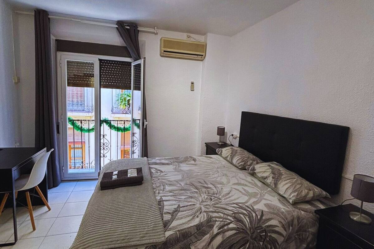 A bedroom with a bed, a desk, a chair, a nightstand, and an air conditioning unit.