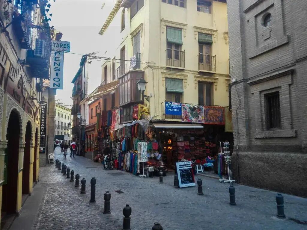Une rue pavée bordée de boutiques et d’immeubles, où l’on aperçoit une enseigne indiquant la présence d’une pension et une vitrine annonçant une vente.