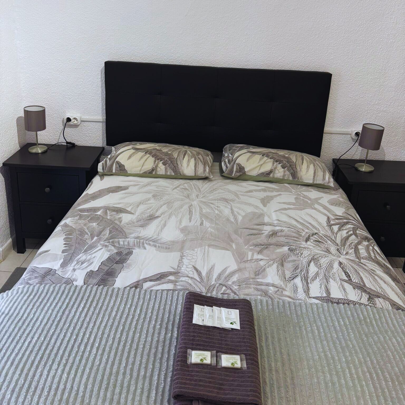 Una cama bien tendida con un edredón estampado con hojas tropicales, flanqueada por dos mesitas de noche con lámparas, frente a una pared con textura.