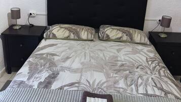 Una cama bien tendida con un edredón estampado con hojas tropicales, flanqueada por dos mesitas de noche con lámparas, frente a una pared con textura.