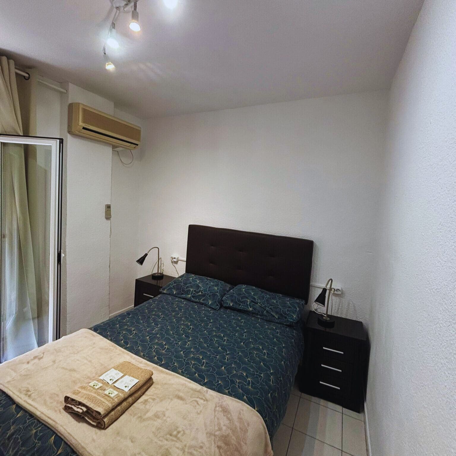 A bedroom with a bed, bedside tables, a nightstand, a window with curtains, and an air conditioning unit.