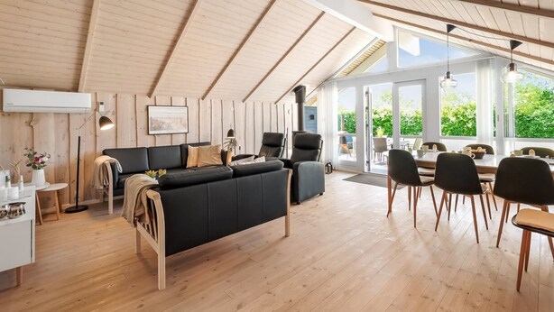 Un vaste espace de vie comprenant un canapé noir, une table à manger et des chaises, un plancher en bois et un plafond en pente avec des poutres apparentes.