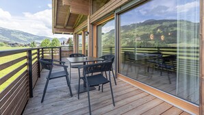 Een houten terras met een tafel en stoelen, grote glasramen en uitzicht op groene heuvels.