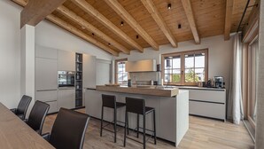 Een moderne keuken met houten balken, een centraal eiland met barkrukken en een eethoek met een lange tafel.