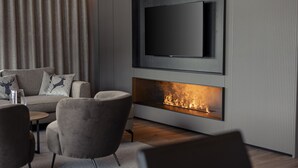 Een moderne woonkamer met een flatscreen televisie aan de muur, een open haard eronder en een bank met decoratieve kussens.