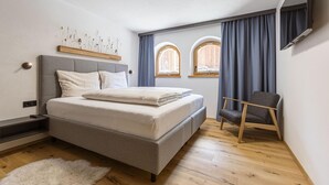 A modern bedroom with a bed, a chair, and two windows with curtains. - Apartment in Tyrol on the ski-slope (Lermoos)