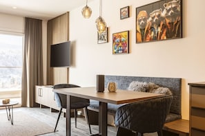 A modern living room with a wooden dining table, black chairs, a grey sofa, and a flat-screen TV mounted on the wall.