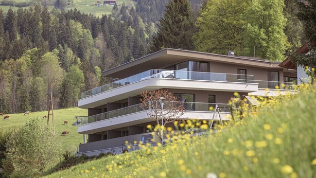 A modern multi-story building with balconies overlooking a green landscape with trees and a field.