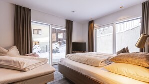 A hotel room with two beds, a TV, and large windows offering a view of a snowy outdoor area.