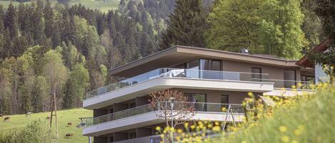 A modern multi-story building with balconies overlooking a green landscape with trees and a field.