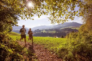 Two people running on a trail surrounded by greenery and mountains.