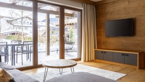 A modern living room with a large sliding glass door leading to an outdoor patio, a flat-screen TV mounted on a wooden wall, and a small round table. - Premium apartment with garden (No Deposit) (Ehrwald)