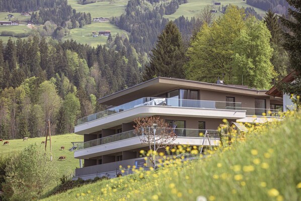 A modern multi-story building with balconies overlooking a green landscape with trees and a field.