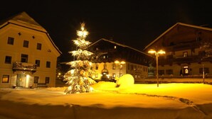 A snow-covered courtyard with a lit Christmas tree and a snowman, surrounded by buildings with balconies and windows.