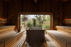 A sauna with wooden benches and a central stone heating area, surrounded by a wooden interior and a view of a forested area. - Spacious apartment with swimming pool (No Deposit) (Ehrwald)