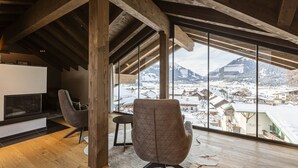 Um quarto com pé-direito alto, vigas de madeira, lareira e uma grande janela com vista para uma aldeia coberta de neve.