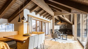 Um amplo loft com teto alto, vigas de madeira, área de jantar com uma mesa comprida e cadeiras, espaço de estar aconchegante com sofás e grandes janelas com vista.
