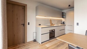 A modern kitchen with a wooden door, white cabinets, a built-in oven, and a wooden dining table. - Beautiful apartment in Tyrol with terrace (Westendorf)