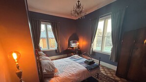 Une chambre à coucher avec un grand lit, un lustre, un bureau avec une lampe et une vue sur l’extérieur par les fenêtres.