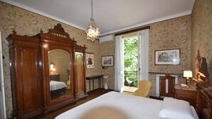 Une chambre avec un grand lit, une armoire en bois et un lustre. Une fenêtre offre une vue sur la verdure environnante.