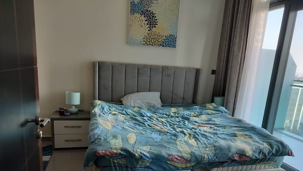A bedroom with a large bed, a nightstand, a lamp, and a window with curtains. - 917a Merano -cozy 1 BHK Apartment (Dubai)