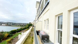 Un balcon donnant sur un plan d’eau et sur de la verdure, un bâtiment blanc avec une fenêtre, et un petit coin salon avec des coussins et une bouteille de vin.