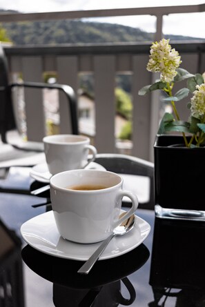 Duas chávenas de café brancas sobre uma mesa preta, com uma colher, uma planta em vaso com flores brancas e uma vista de montanha ao fundo.