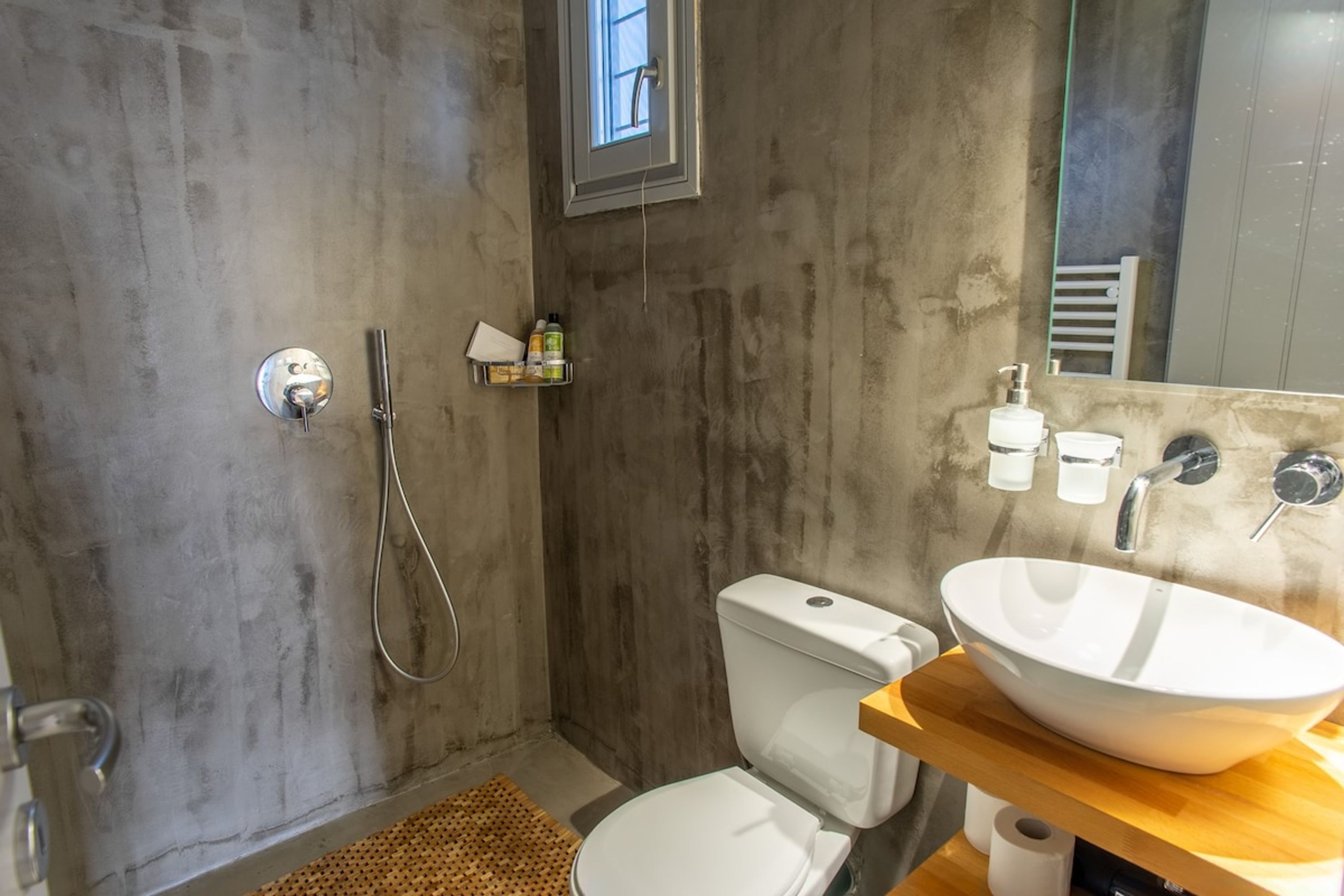 Un baño con pared de concreto, inodoro blanco, tocador de madera con un lavamanos grande y blanco, ducha con cabezal de ducha manual y un estante pequeño con artículos de tocador.